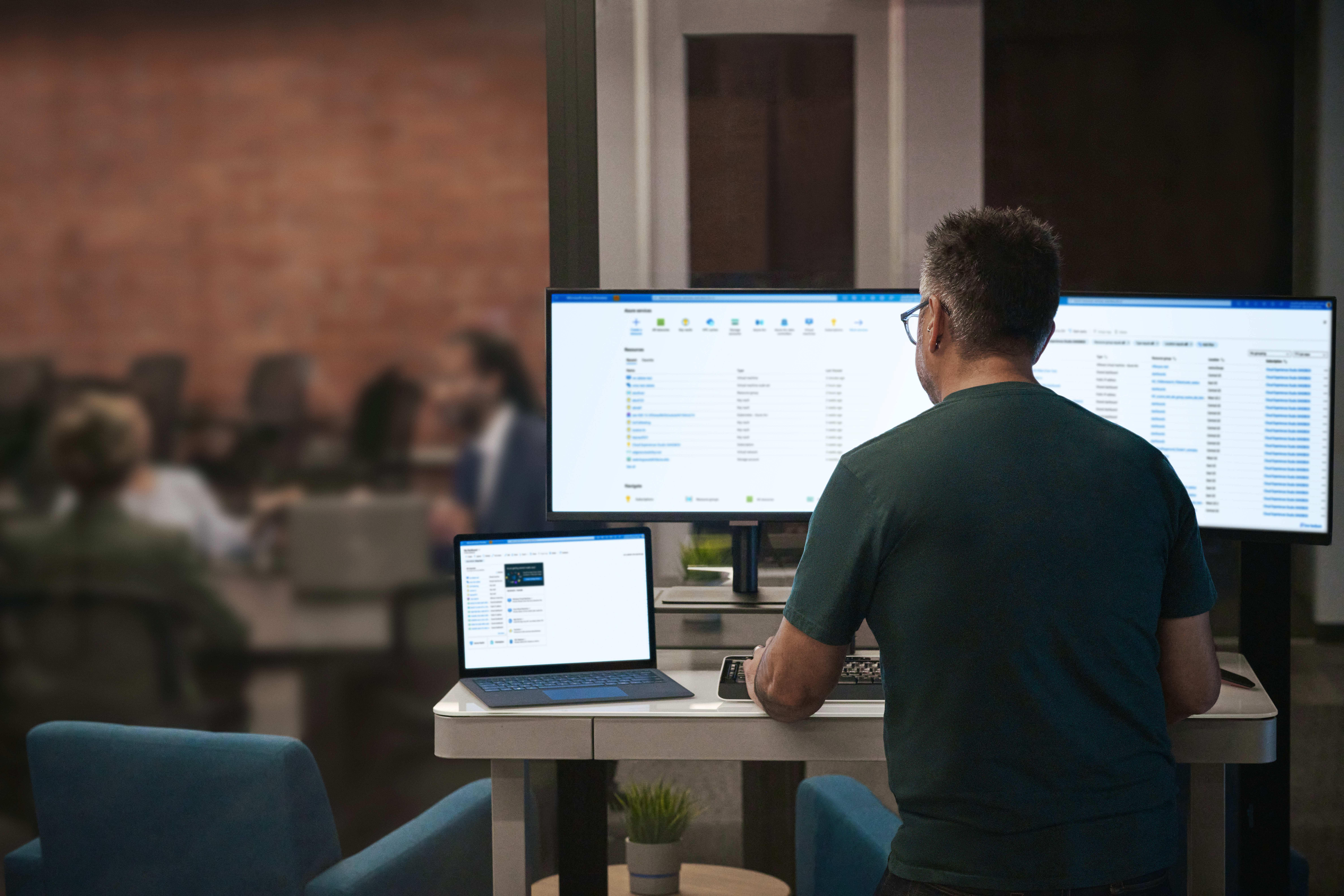 Na imagem, um homem está de costas, trabalhando em dois computadores e um notebook.