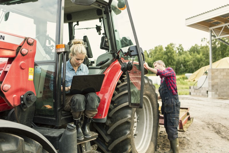 Una mujer trabaja en una laptop sentada en un tractor mientras un hombre revisa el vehículo