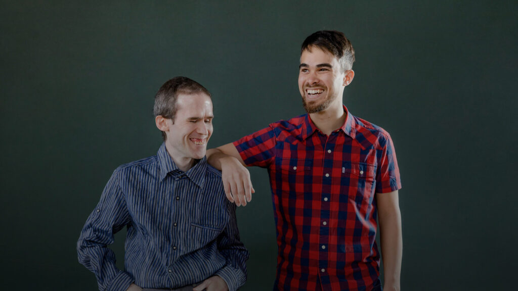 Dos hombres sonrientes, uno con el brazo alrededor del otro, de pie sobre un fondo verde oscuro. Llevan camisas informales.