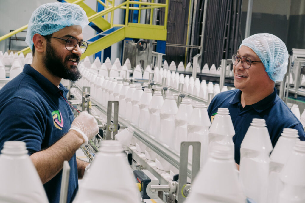 La imagen muestra a dos personas con gorros azules, camisas azules y guantes blancos, de pie en un entorno de fábrica. Están rodeadas por filas de botellas de plástico blancas en un sistema de cinta transportadora. El fondo incluye equipo industrial, barandillas metálicas y una escalera amarilla. El ambiente parece estar limpio y organizado, lo que indica que se trata de una instalación de fabricación o envasado.