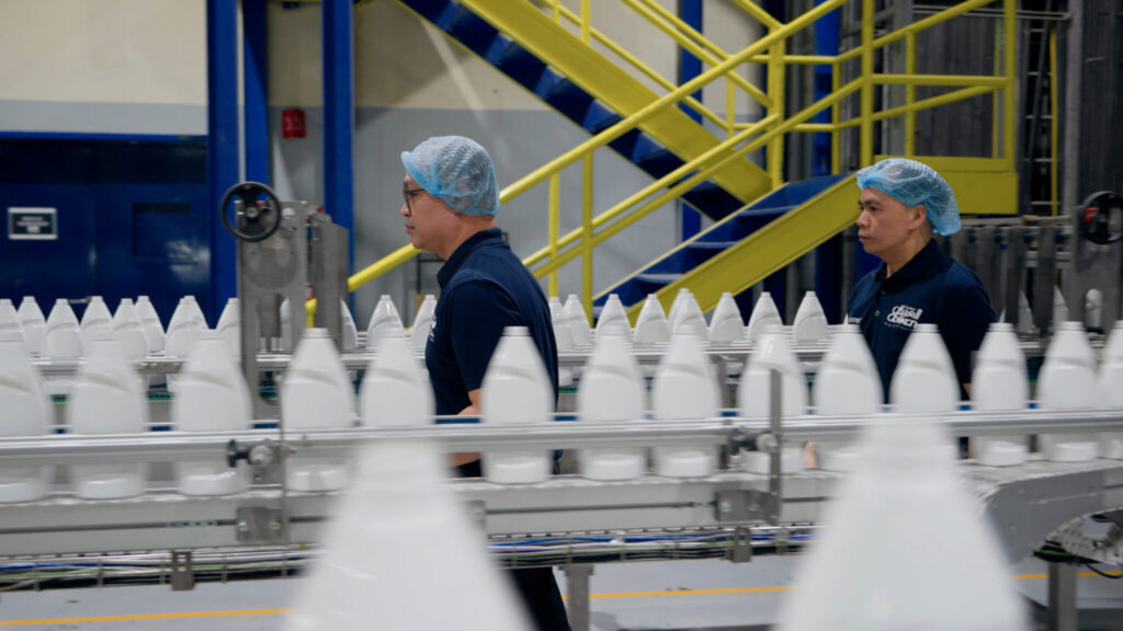La imagen muestra un entorno de fábrica con dos personas que llevan gorros azules y camisas azul oscuro. Están de pie junto a una cinta transportadora que transporta botellas de plástico blancas. En el fondo, hay una escalera amarilla con barandillas azules y las paredes están pintadas de azul y gris. Alrededor de la cinta transportadora se pueden ver varias piezas de equipo y maquinaria industrial.