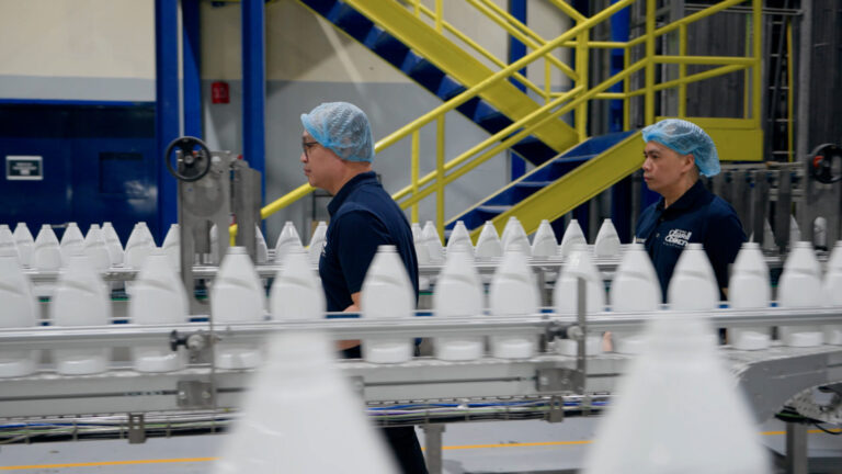 La imagen muestra un entorno de fábrica con dos personas que llevan gorros azules y camisas azul oscuro. Están de pie junto a una cinta transportadora que transporta botellas de plástico blancas. En el fondo, hay una escalera amarilla con barandillas azules y las paredes están pintadas de azul y gris. Alrededor de la cinta transportadora se pueden ver varias piezas de equipo y maquinaria industrial.