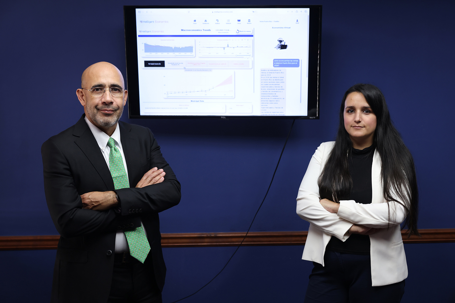 A man and a woman stand in front of a blue wall with a computer monitor between them