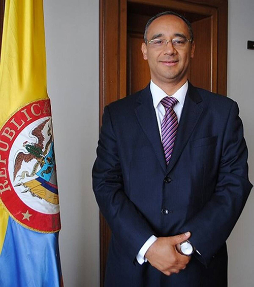Um homem de terno está ao lado da bandeira colombiana com as mãos cruzadas na frente do corpo