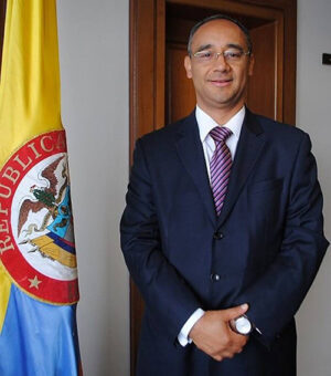 Un hombre con traje está de pie junto a la bandera colombiana con las manos cruzadas frente a él.